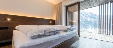 A modern bedroom with a wooden headboard, a large bed with white linens, and a view of mountains through a sliding glass door.