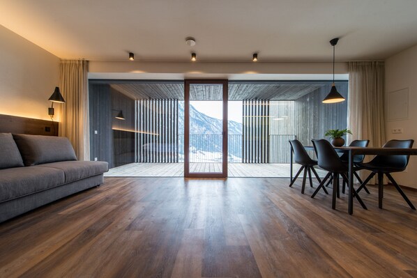 A modern living room with a sofa, dining table, chairs, and a view through a glass door to a balcony. - Mountain Residence Montana (Valle di Casies)