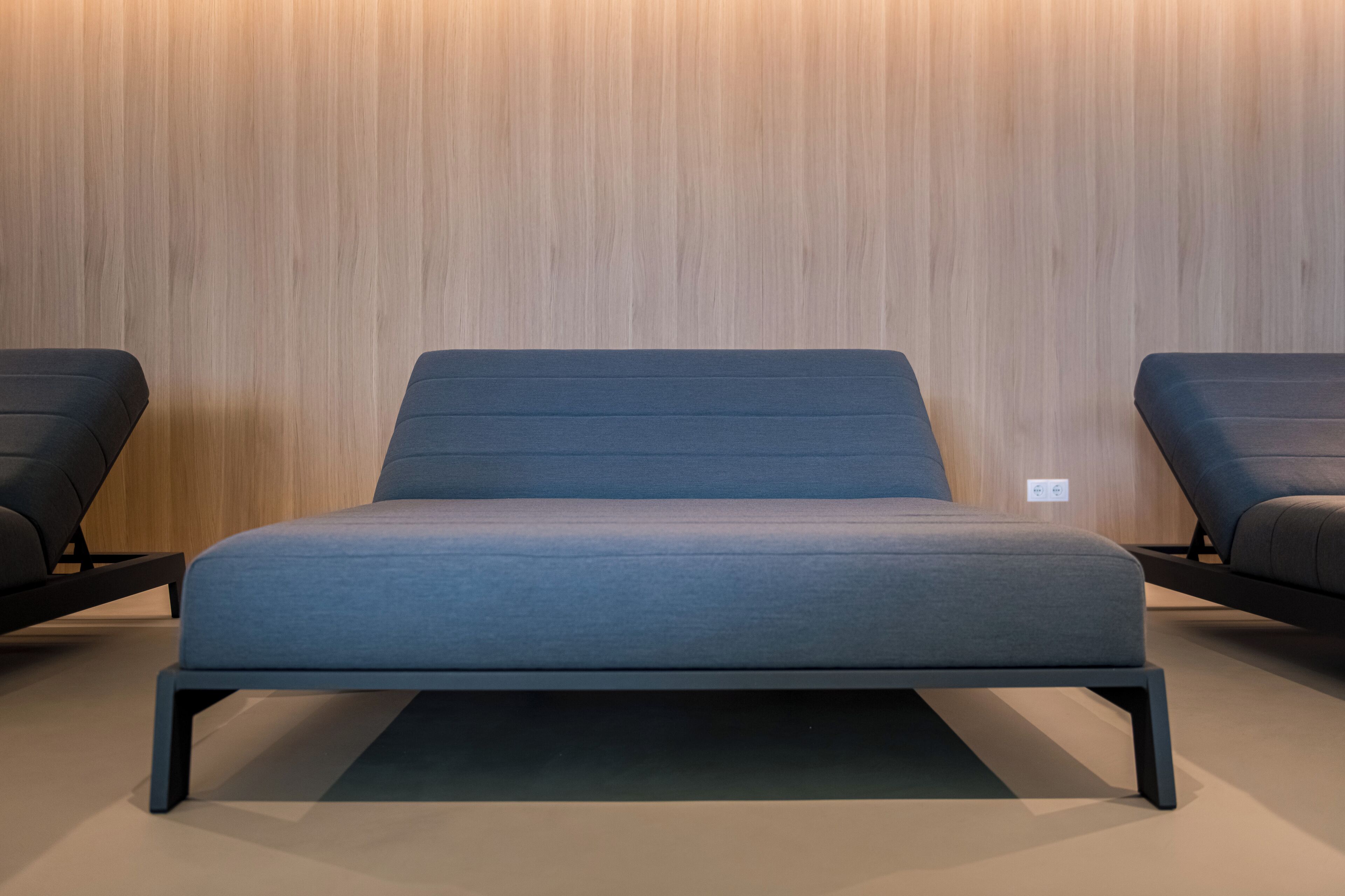 Un lit moderne avec une tête de lit et un pied de lit bleu foncé, placé contre un mur en bois sur lequel on peut voir une prise de courant.