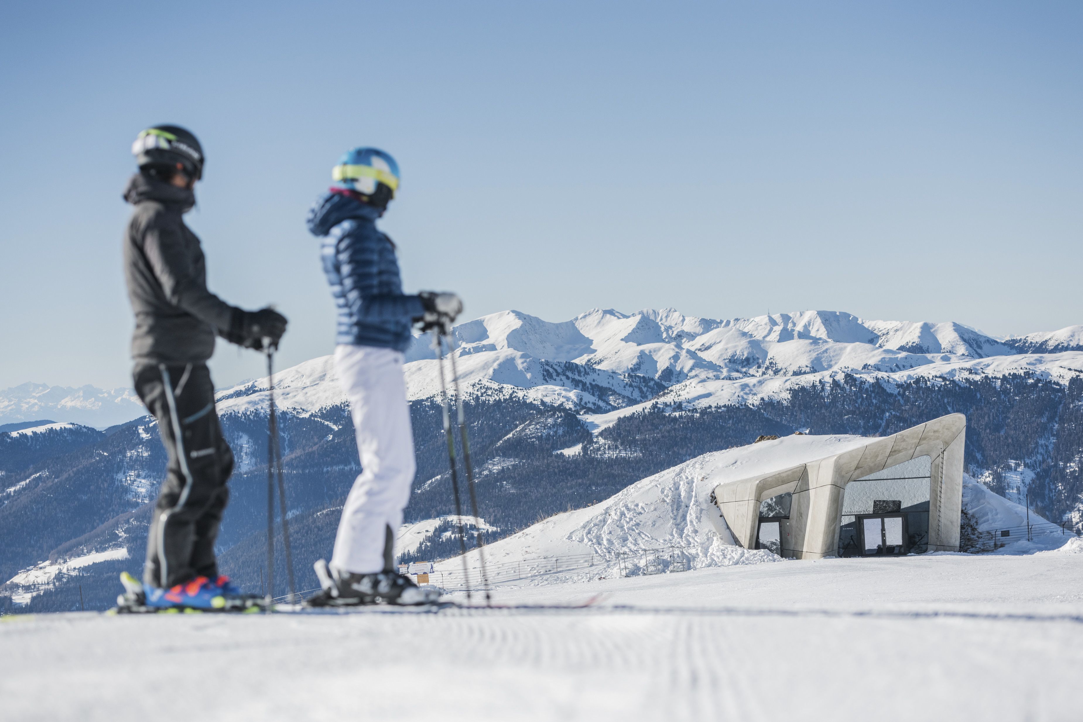 Eine Skipiste mit zwei Skifahrern, einem verschneiten Gebirge und einer Betonstruktur im Hintergrund.