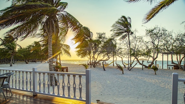 Eine Holzterrasse mit weißer Brüstung blickt auf einen Strand mit Palmen und einen Sonnenuntergang.