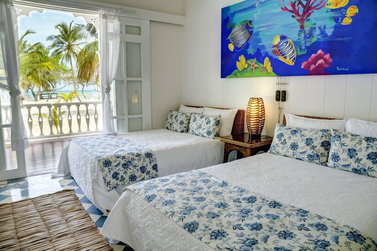Ein Hotelzimmer mit zwei Betten, einem großen Fenster mit Blick auf Palmen und das Meer sowie einem farbenfrohen Fischgemälde an der Wand.