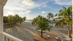 Blick auf einen Strand von einer überdachten Terrasse mit Holzmöbeln und Palmen.