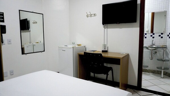 A hotel room with a bed, a desk with a chair, a refrigerator, a television, and a bathroom with a sink and mirror.