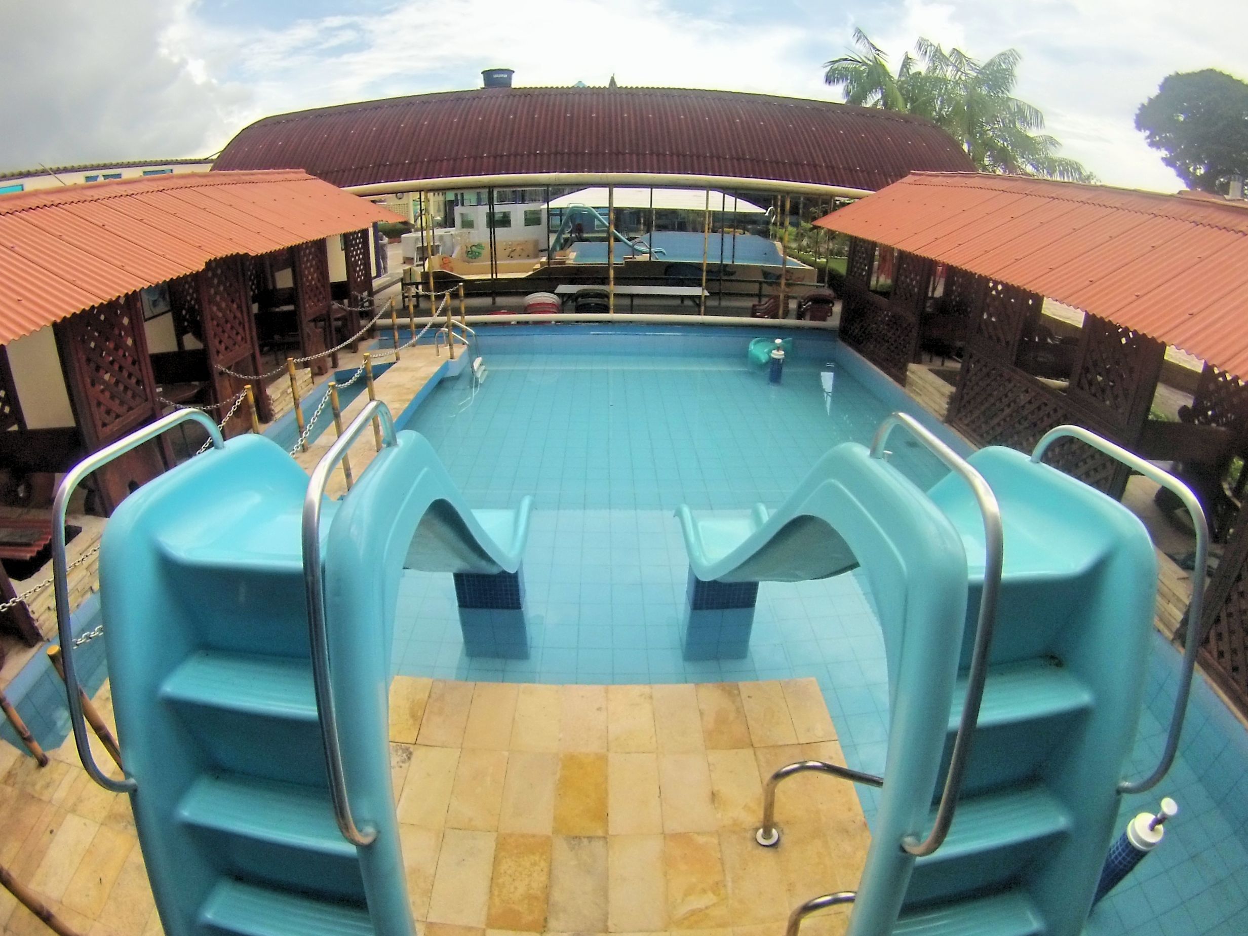 Una piscina con borde de baldosas, barandillas de metal y una zona cubierta con techo rojo.