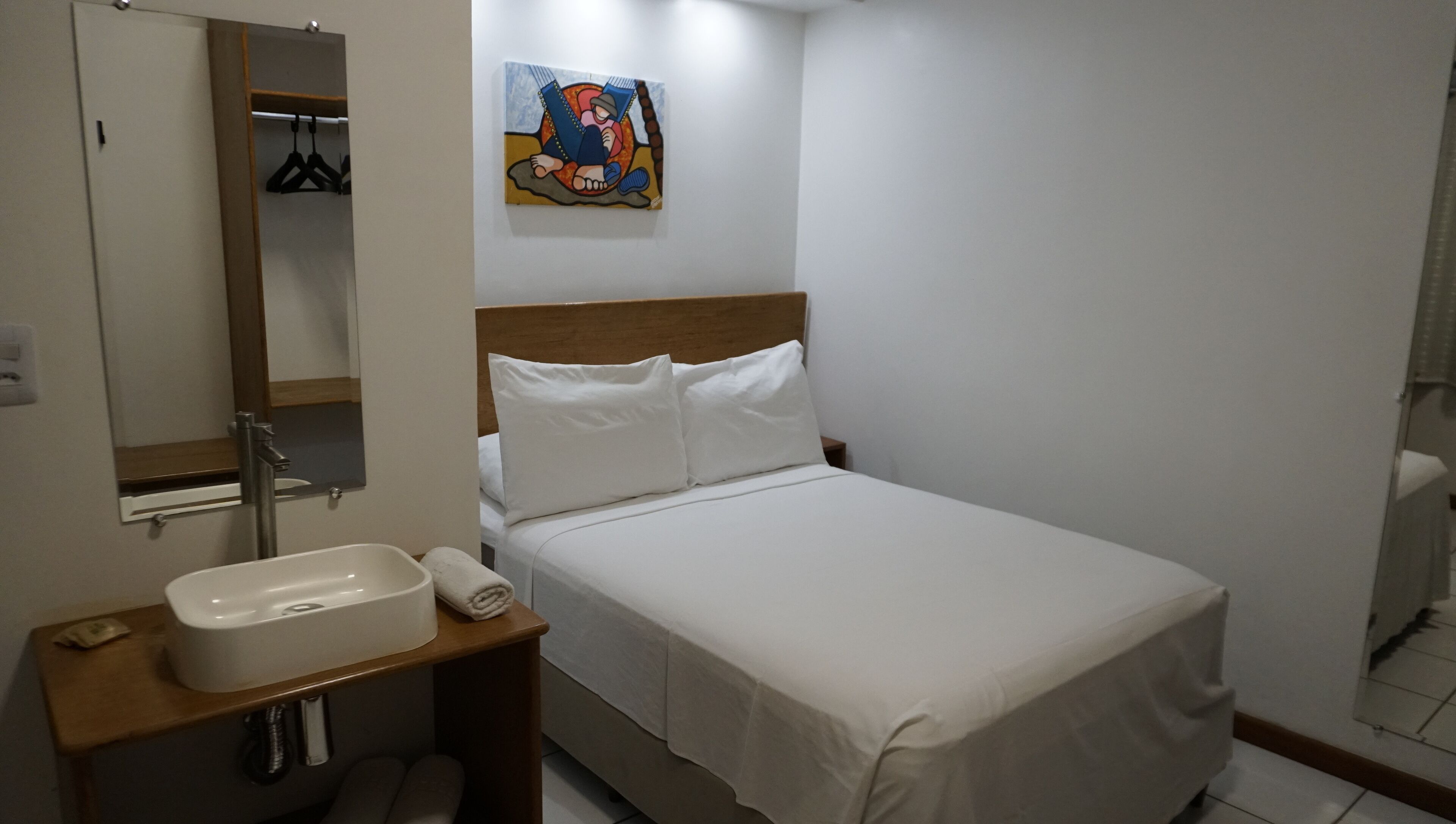 A hotel room with a bed, a wooden nightstand with a white sink, a towel, and a painting on the wall.