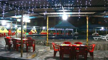 Un patio al aire libre con mesas, sillas, piscina y luces colgantes.