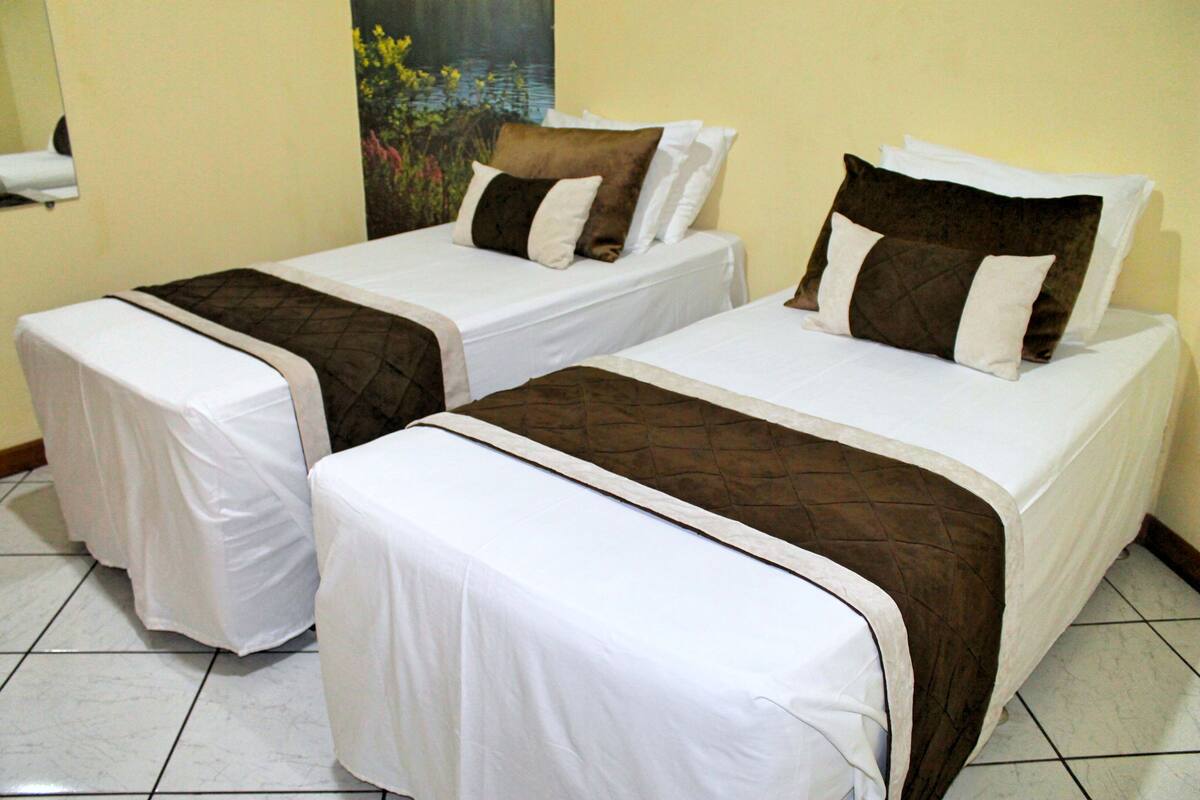 Two single beds with white and brown bedding, a painting on the wall, and a tiled floor.