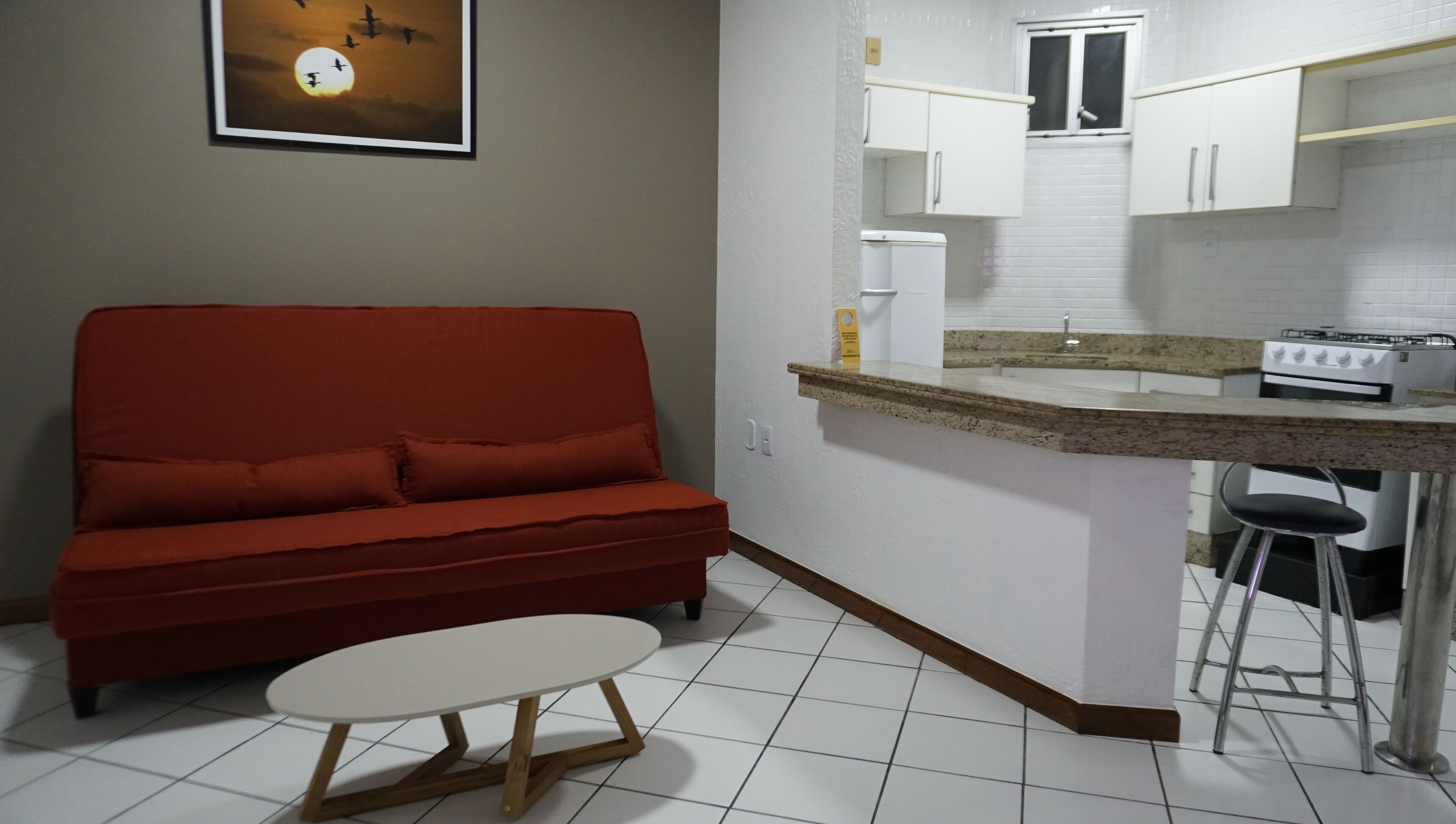 Un espacio habitable compacto con un sofá rojo, una pequeña mesa redonda, una zona de cocina con armarios blancos, una estufa a gas y suelo de baldosas.
