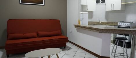 Un espacio habitable compacto con un sofá rojo, una pequeña mesa redonda, una zona de cocina con armarios blancos, una estufa a gas y suelo de baldosas.
