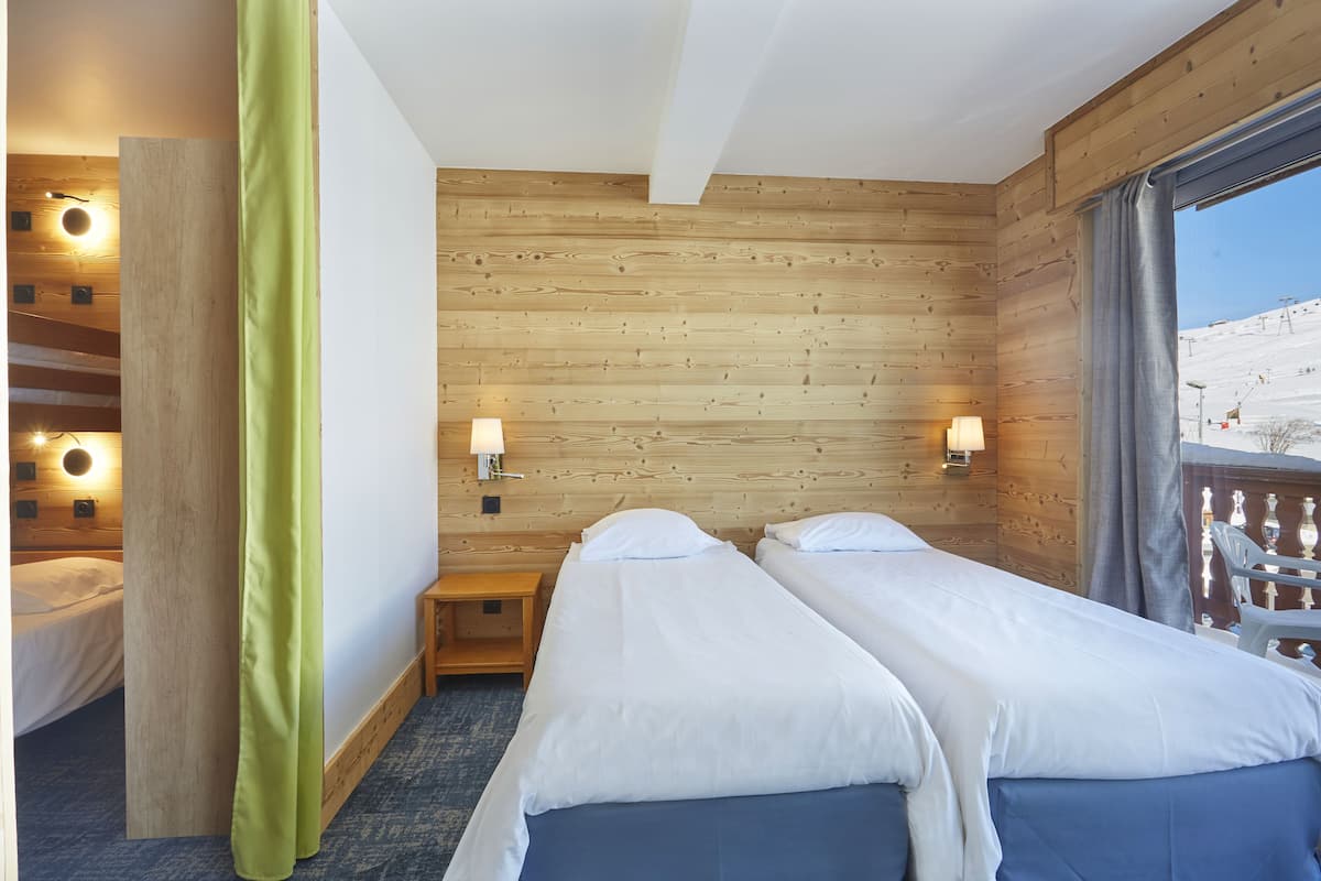 A hotel room with two beds, wooden walls, a window with a view of a ski slope, and wall-mounted lights.