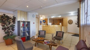 A reception area with a wooden reception desk, two grey armchairs, a coffee table with magazines and a basket, a large potted plant, and a wall clock.