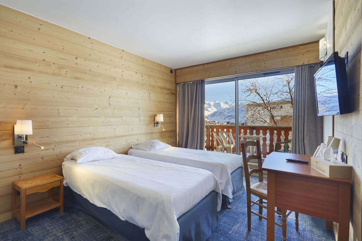 A hotel room with two beds, a desk, a chair, a TV, and a window with a view of mountains.