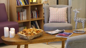 A cozy living room with a wooden bookshelf, a grey armchair, a purple sofa, a wooden coffee table with a basket of pastries, and a decorative reindeer.