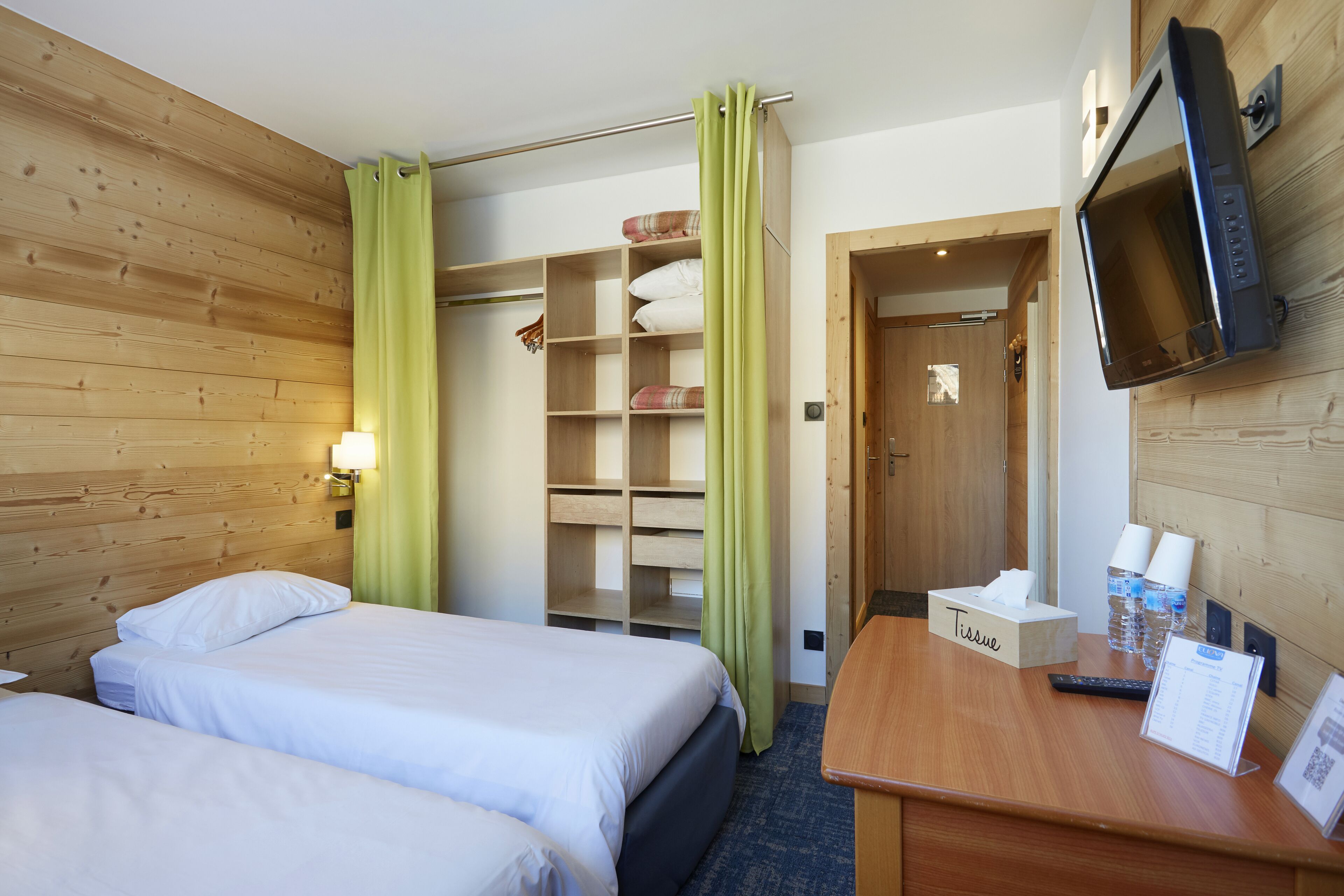 Hotellihuone, jossa on kaksi sänkyä, työpöytä paperipyyhekotelolla, televisio ja puinen vaatekaappi.