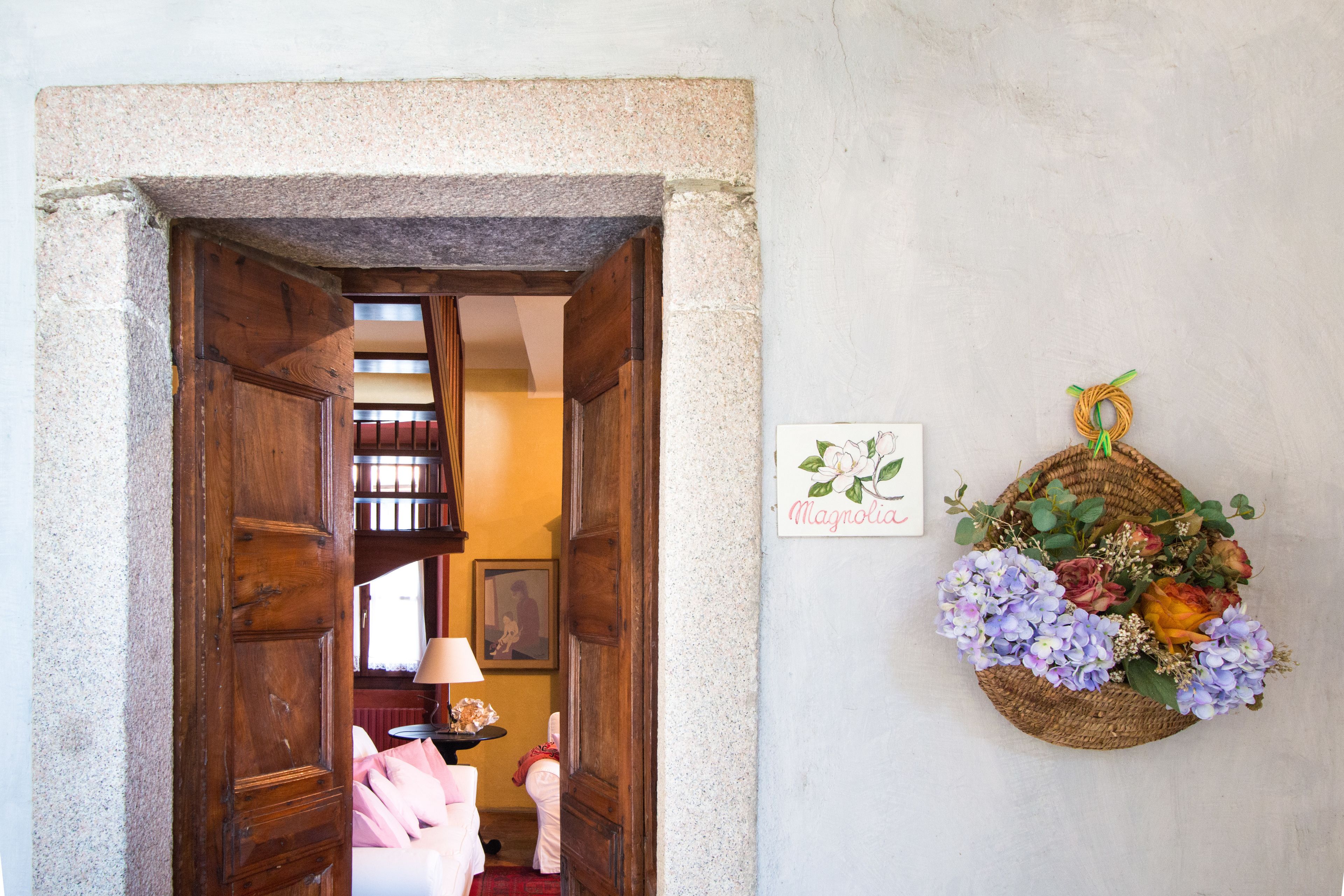 Una porta d'ingresso che dà su una stanza con un letto e una lampada, un cartello appeso al muro e un cesto di vimini con fiori.