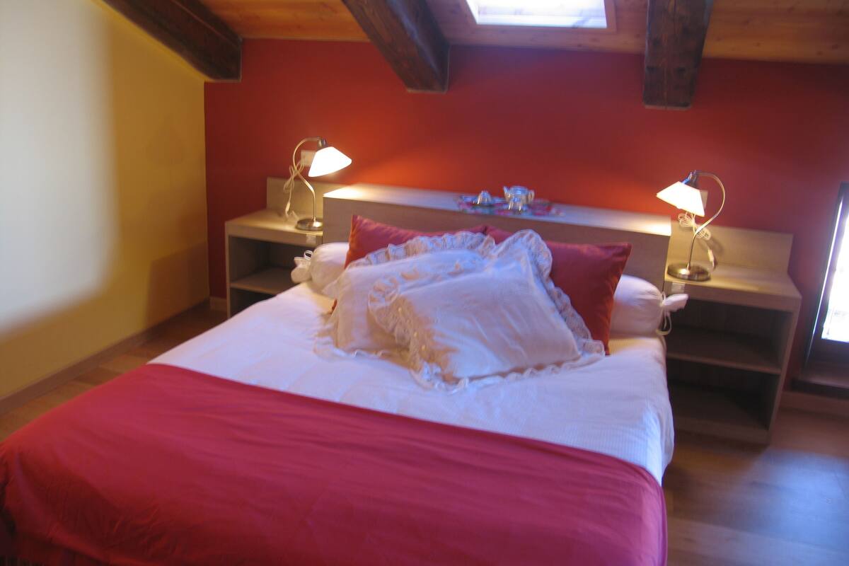 A bedroom with a large bed, two bedside tables with lamps, a wooden ceiling, and a red wall.