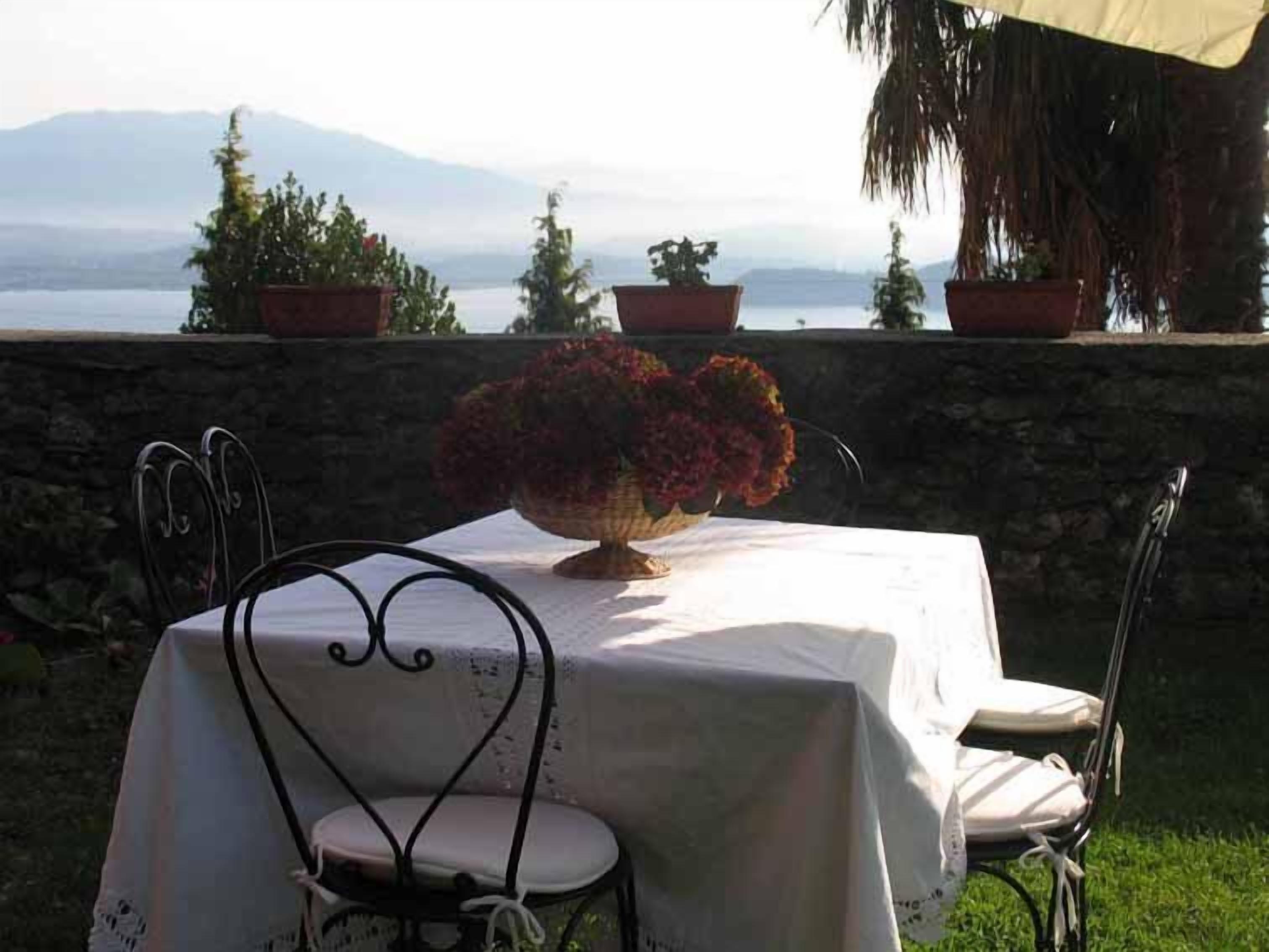A table with a white tablecloth, a vase with flowers, and chairs. There are potted plants on a stone wall and a view of a body of water and mountains in the background.