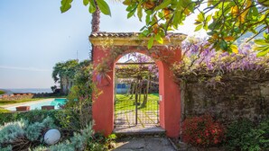 Un chemin de pierres mène à une arche rouge surmontée d’un portail en métal, le tout étant entouré d’une végétation luxuriante et de plantes en fleurs.