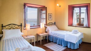 Une chambre avec deux lits, une fenêtre avec des rideaux, une table de chevet avec une lampe et un tableau encadré au mur.