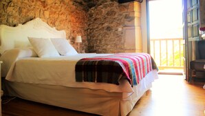 Un dormitorio con pared de piedra, una cama con ropa de cama blanca, una manta colorida y vistas al exterior a través de un ventanal grande.
