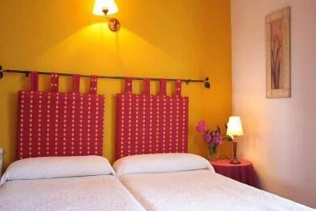 Une chambre avec deux lits, un mur jaune, un rideau rouge et une petite table avec une lampe.