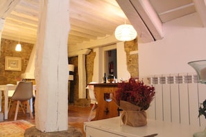 Un interior rústico con vigas de madera a la vista, una pared de piedra, una encimera de madera con una botella y un jarrón con flores rojas.