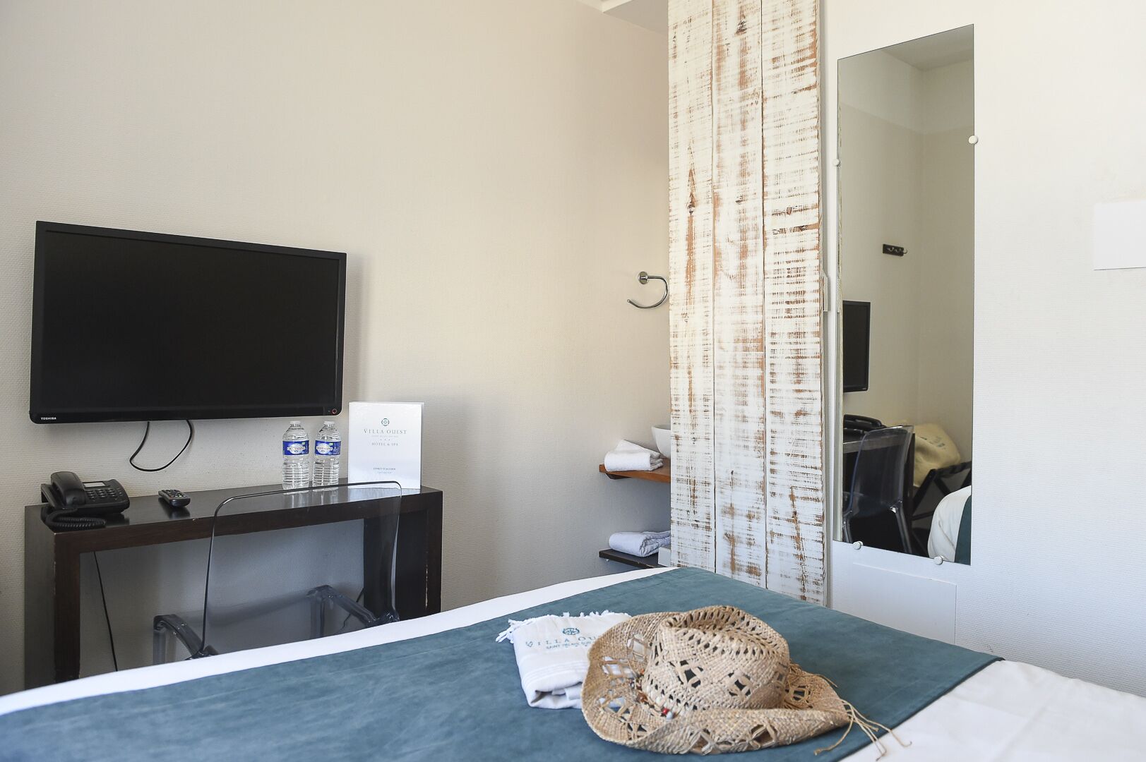 A hotel room with a bed, a desk with a telephone and a sign, a television, a mirror, and a bathroom with a shower curtain.