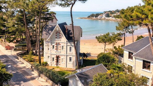 Un bâtiment situé sur la côte, offrant une vue sur la mer, entouré d’arbres et sous un ciel dégagé.