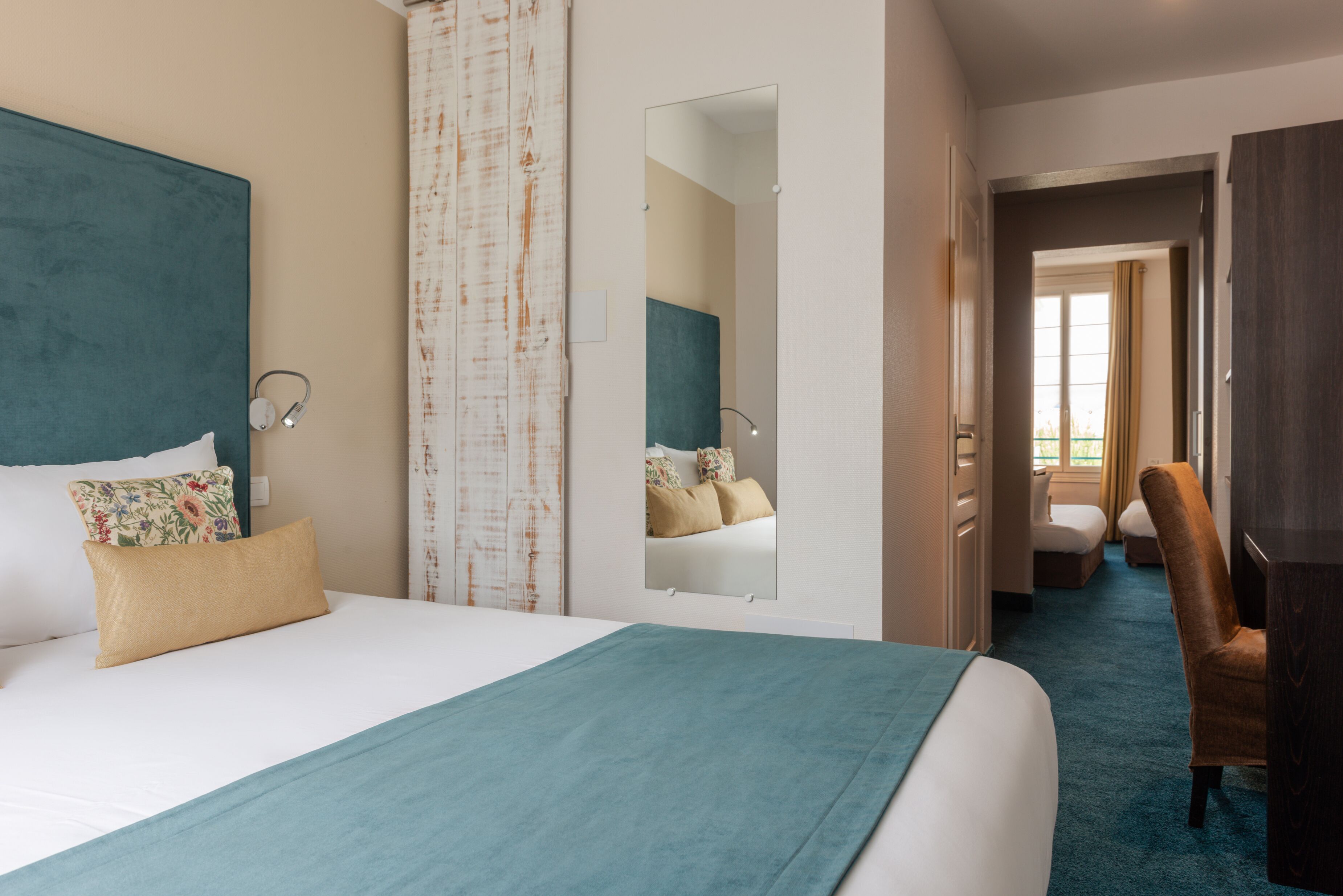 Ein Hotelzimmer mit einem großen Bett, einem Spiegel, einem Sessel und Blick auf ein weiteres Zimmer.