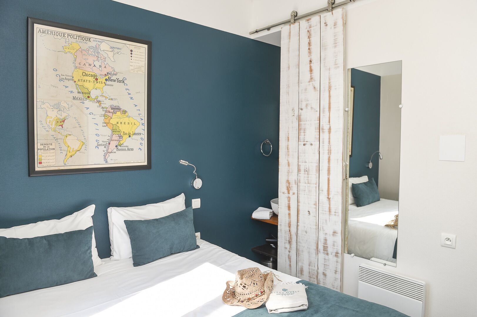 Un dormitorio con cama, almohadas, un mapa en la pared, un espejo y una ventana con cortinas.