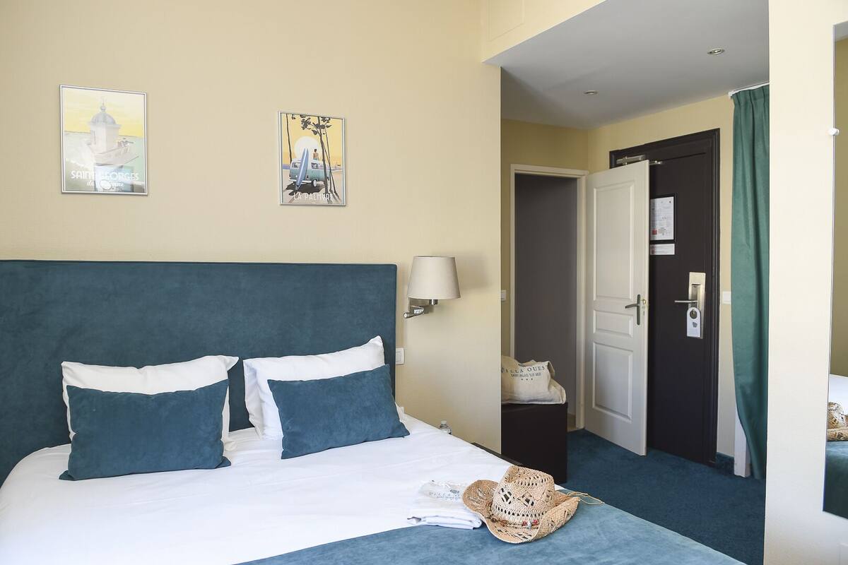 Une chambre d’hôtel avec un lit, des oreillers, un chapeau, une lampe et deux œuvres d’art murales.