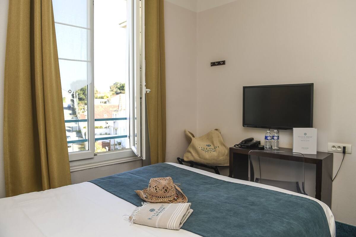 Une chambre d’hôtel avec un lit, un bureau avec un ordinateur, une télévision et une fenêtre avec des rideaux.