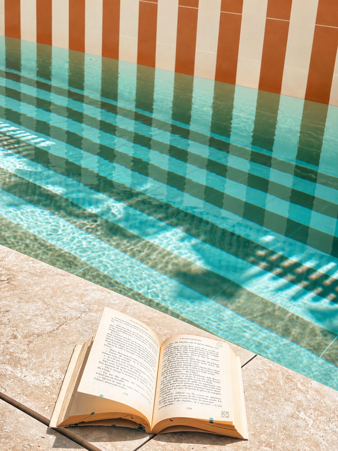 Un livre ouvert sur une surface en pierre, près d’une piscine dont le sol est recouvert de carreaux à motifs.