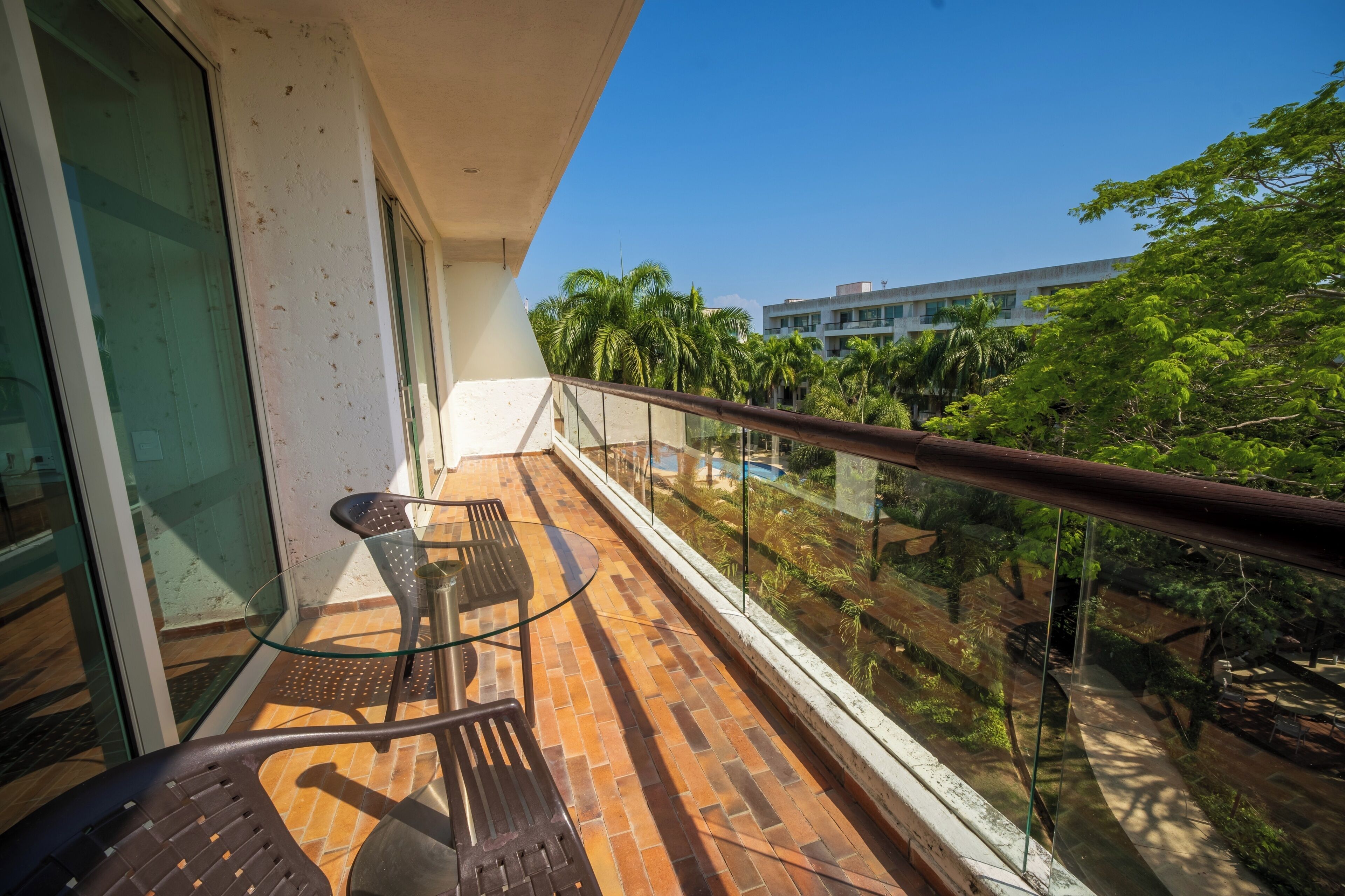 A balcony with a glass railing, a table, and chairs, overlooking a pool and greenery.