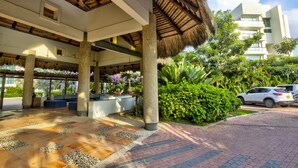 A covered outdoor area with a thatched roof, stone pillars, and a paved walkway leading to a building with large windows and a white facade. - Hotel Estelar Playa Manzanillo (Cartagena)