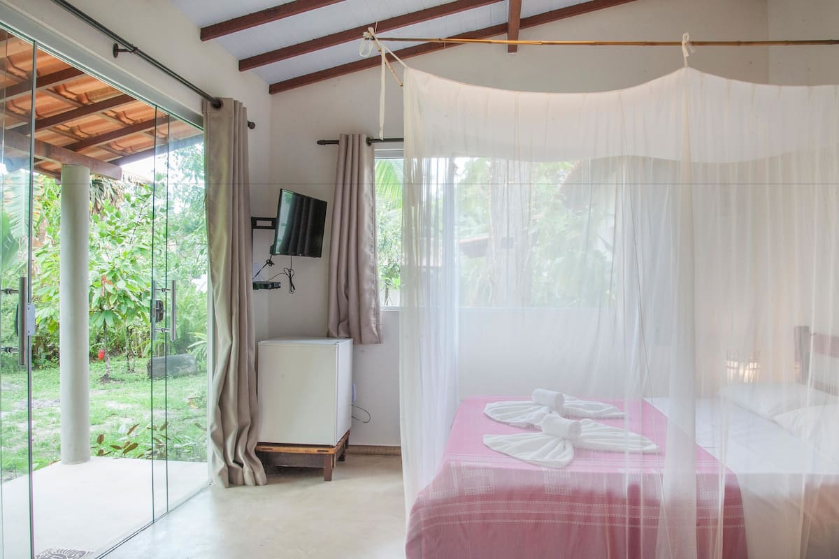 A bedroom with a bed covered by a mosquito net, a television on the wall, and a view of greenery outside.