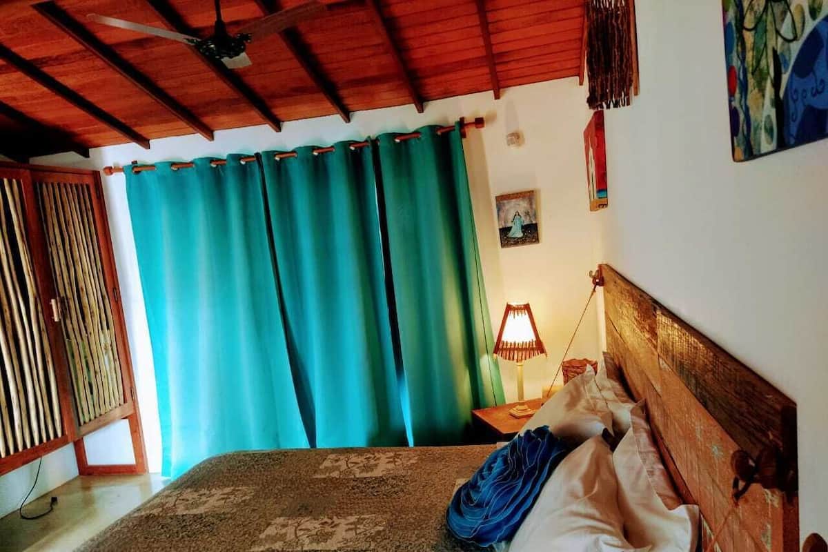 A bedroom with a wooden headboard, a bed with a patterned bedspread, teal curtains, a nightstand with a lamp, and a wall-mounted painting.