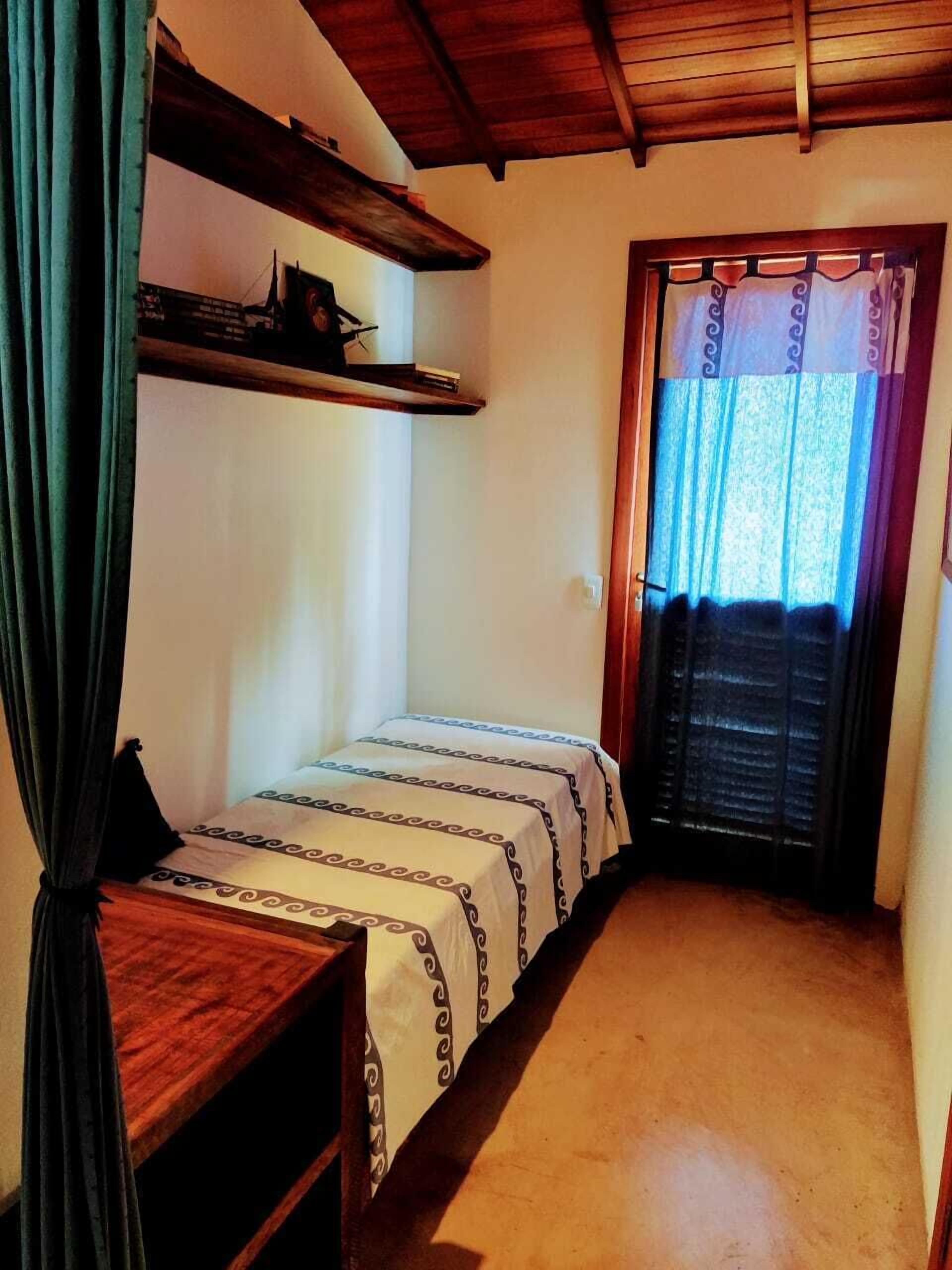 Une petite pièce agréable, avec un lit simple, une étagère en bois et une fenêtre avec des rideaux.