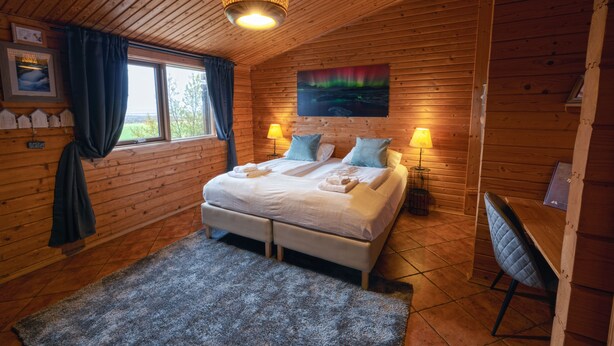 A wooden cabin bedroom with a bed, two bedside lamps, a window with curtains, a painting, and a small table with a chair.