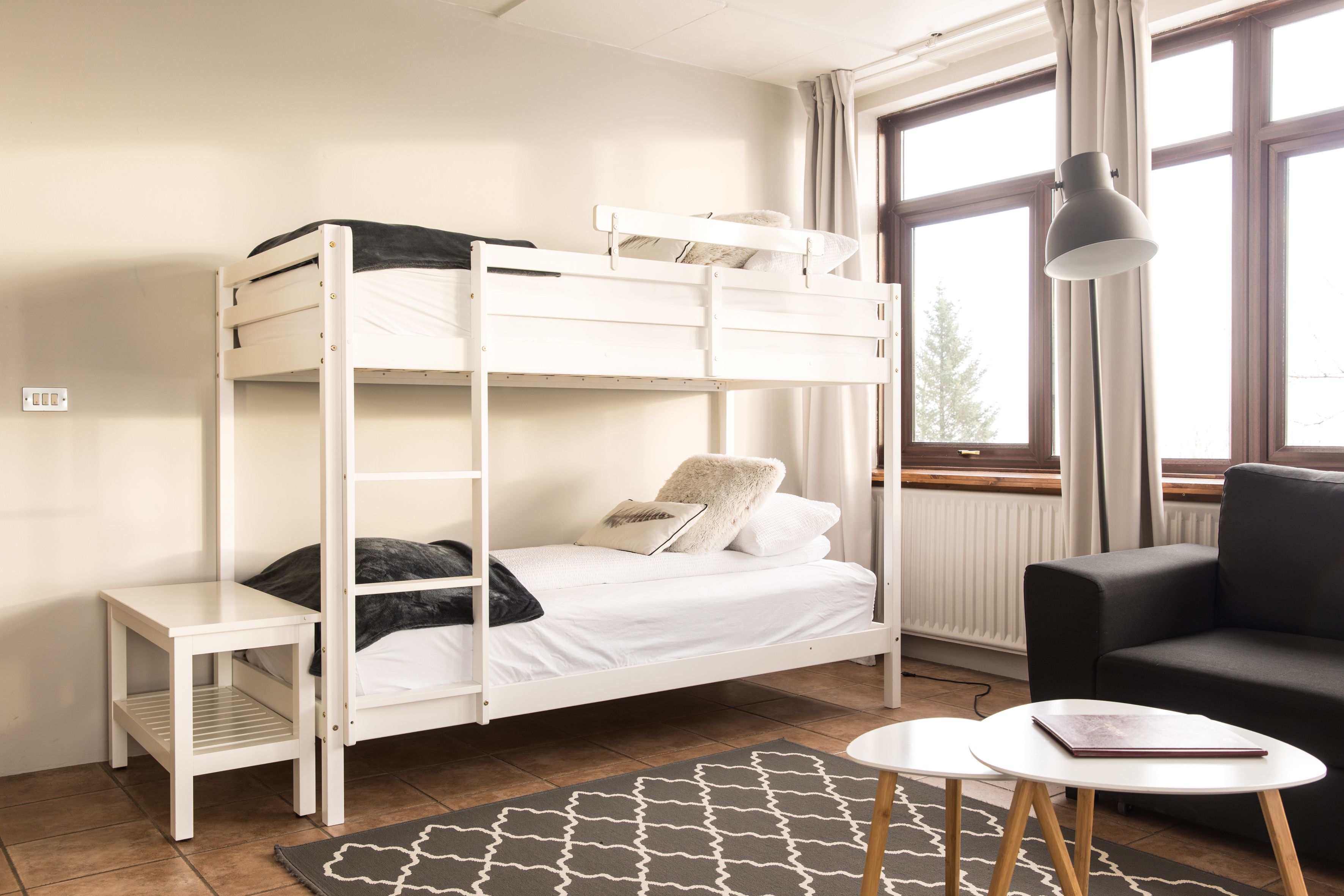 Una cama litera con estructura blanca, una mesita de noche pequeña, un sofá negro y una lámpara.