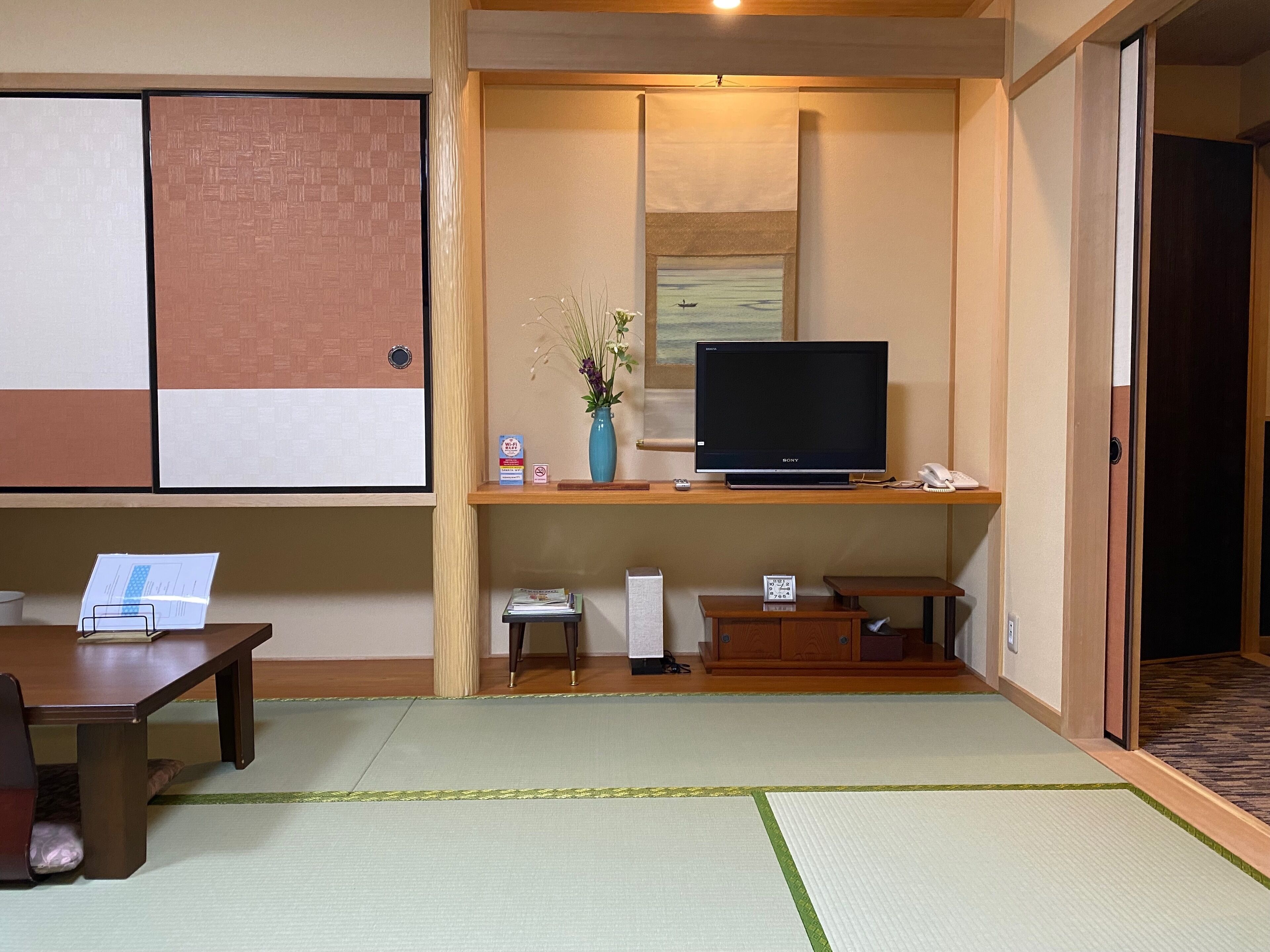 A traditional Japanese room with tatami flooring, a television on a wooden stand, a vase with flowers, and a sliding door.