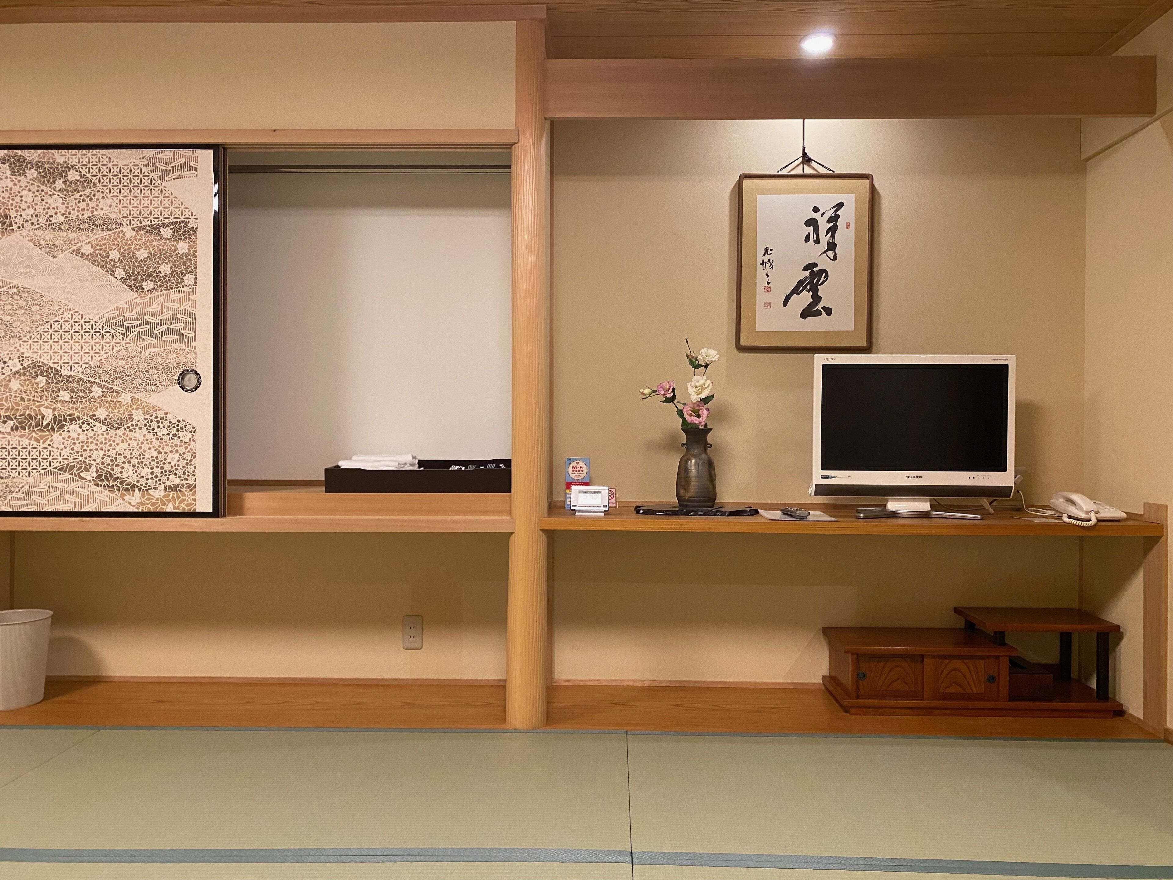 A traditional Japanese room with tatami flooring, a shoji screen, a desk with a computer, a vase with flowers, and a calligraphy piece on the wall.