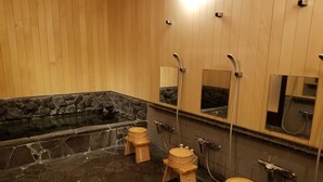 Een badkamer met een stenen vloer, houten krukken, een bad met een stenen wand en een grote spiegel.