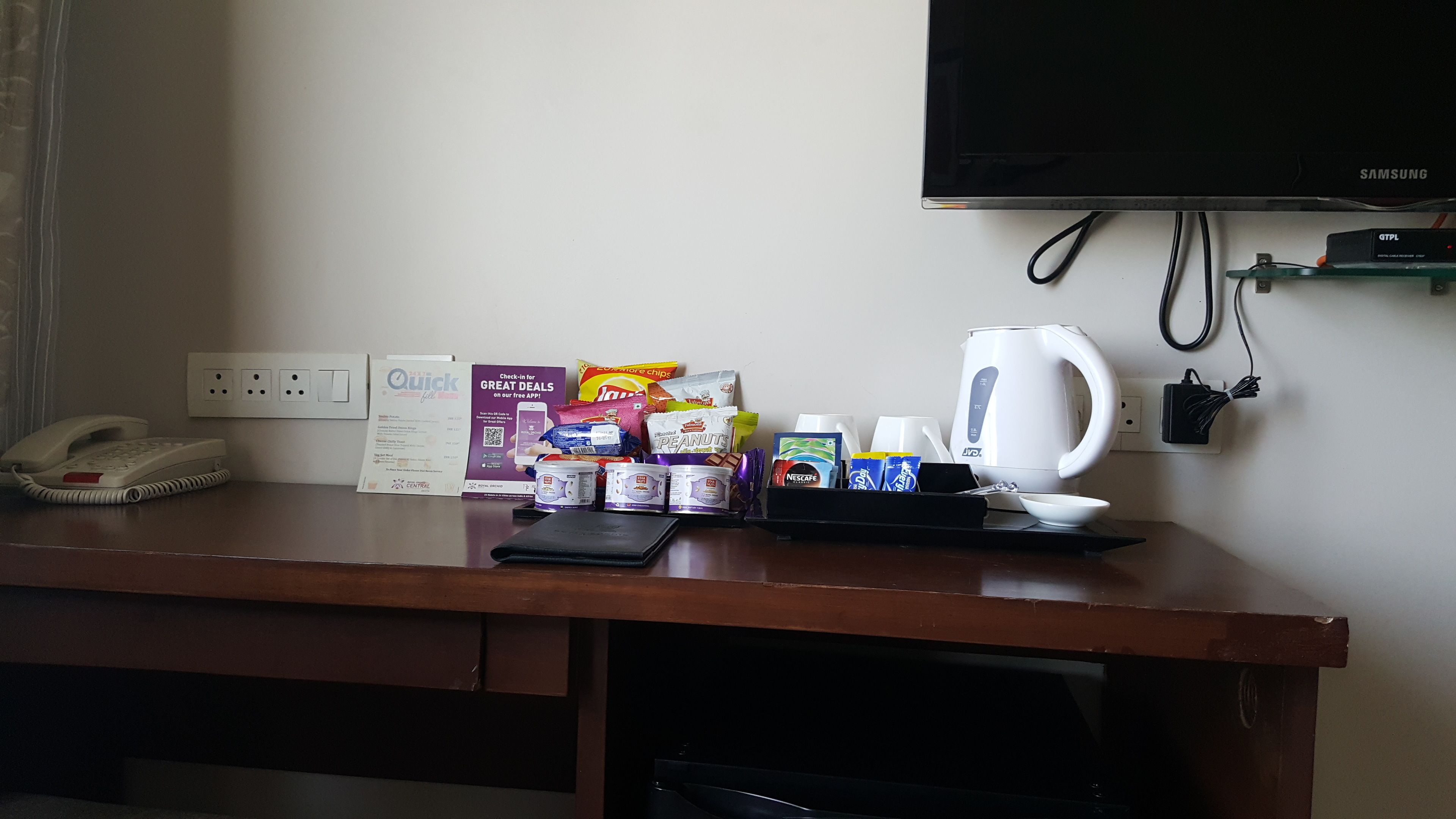 A hotel desk with a phone, a sign for a quick check-in app, snacks, a kettle, and a TV.