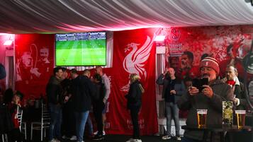A room with a large screen displaying a football match, a red wall with a white logo, and a table with drinks.