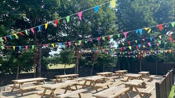 An outdoor area with picnic tables arranged in rows, surrounded by a wooden fence, and decorated with multicolored triangular flags.
