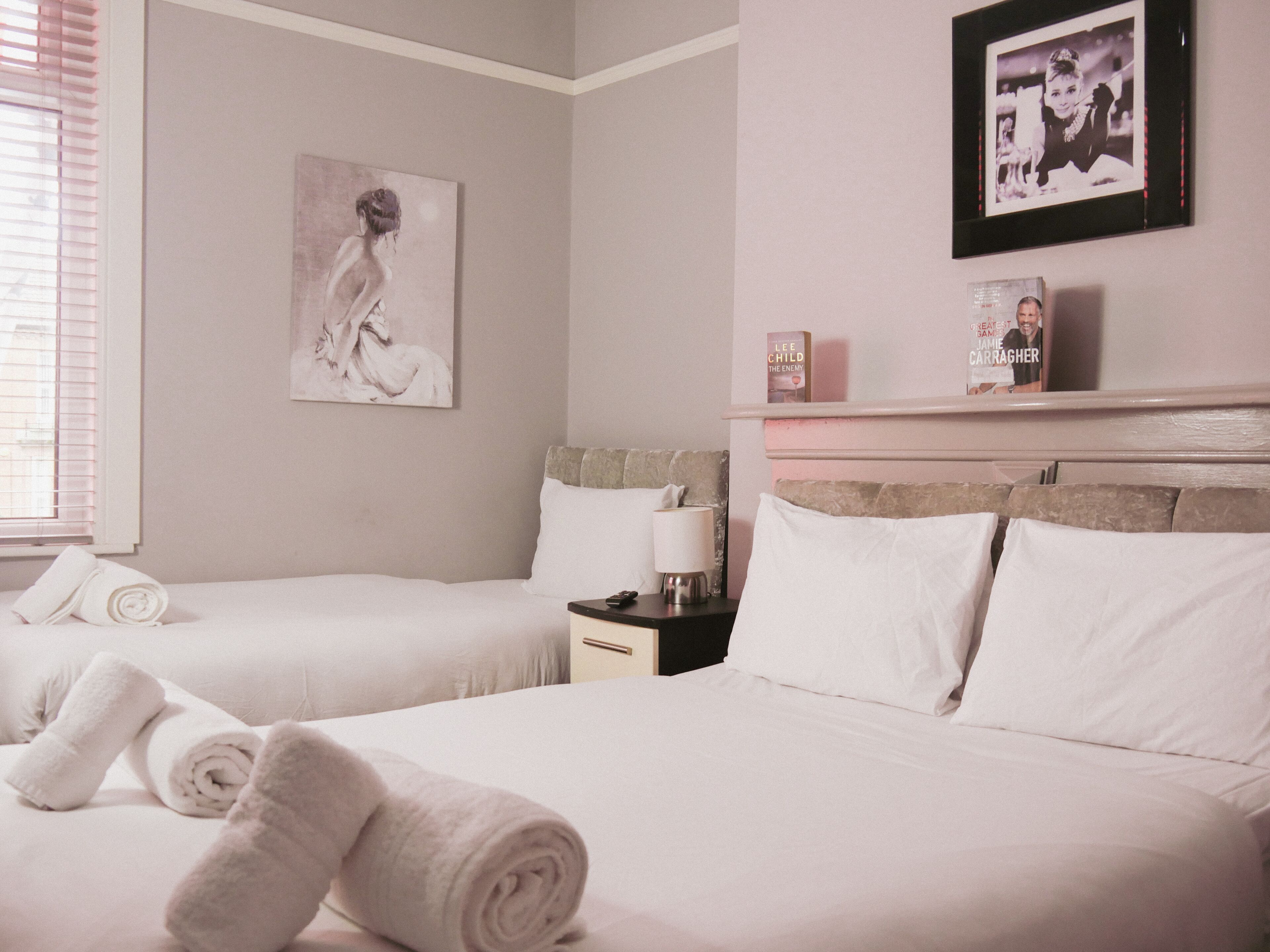 Une chambre d’hôtel avec deux lits, du linge de lit blanc, un tableau au mur et une étagère avec des livres et une photographie encadrée.