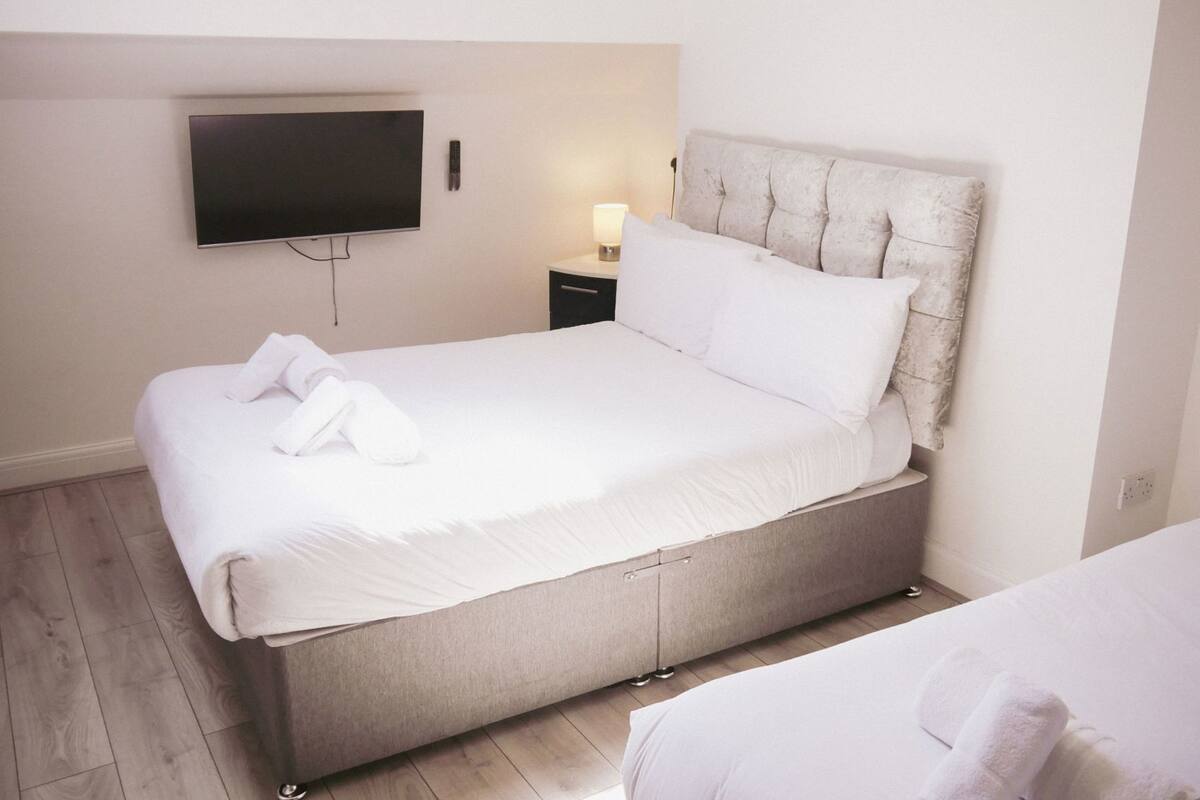 Un lit bien fait, avec du linge de lit blanc et une serviette pliée, une table de chevet avec une lampe et un téléviseur à écran plat fixé au mur.