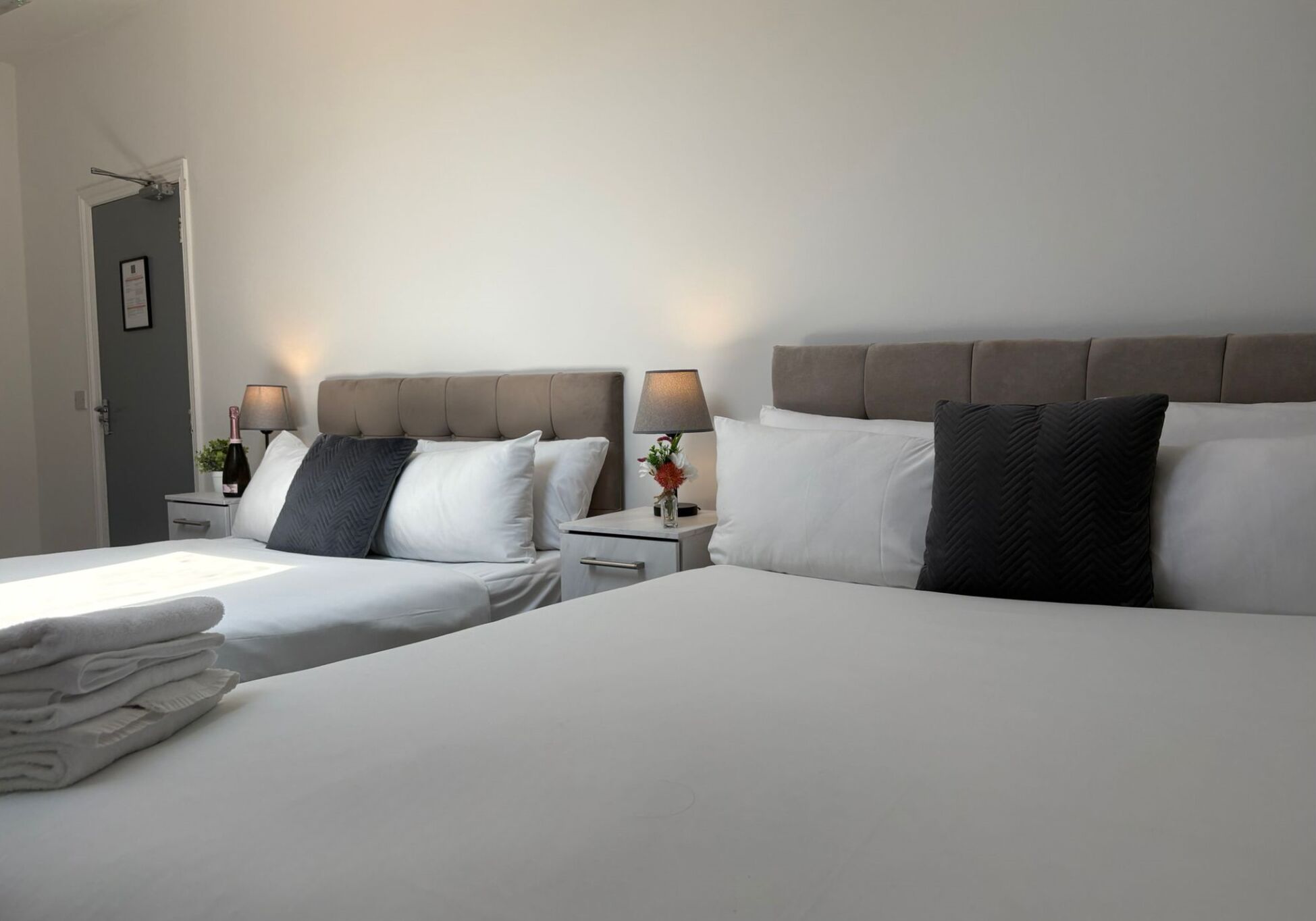 A hotel room with two beds, each with white linens and a grey headboard. There are bedside tables with lamps and a small vase with flowers.